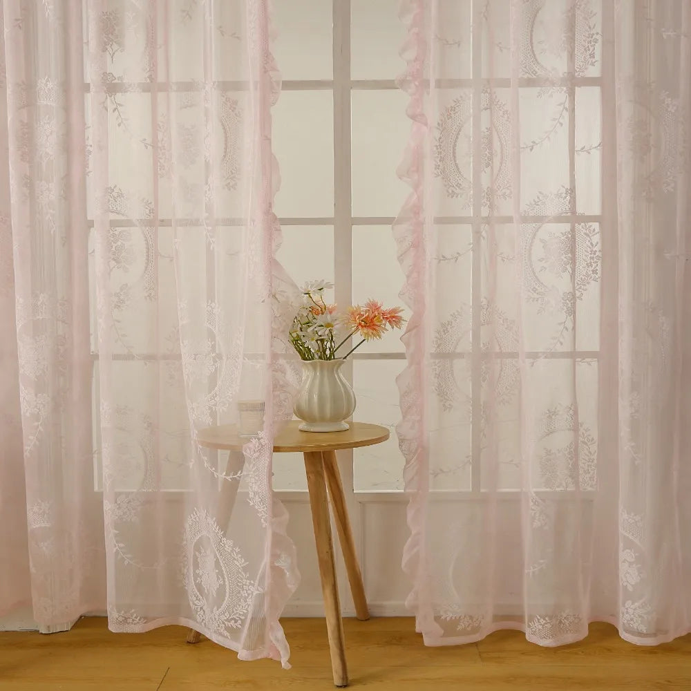 Pink ruffled French lace sheer curtains with floral embroidery hanging in a window.