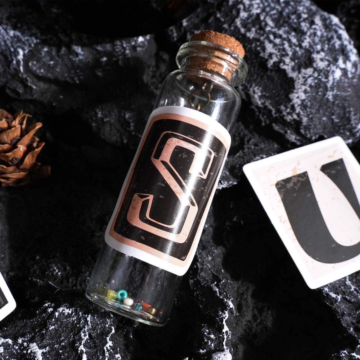 A glass bottle filled with alphabet stickers is displayed on a dark surface next to a white card.