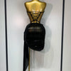 A black, ruched dress is displayed on a gold mannequin, featuring a laced-up bodice and a long black drape.