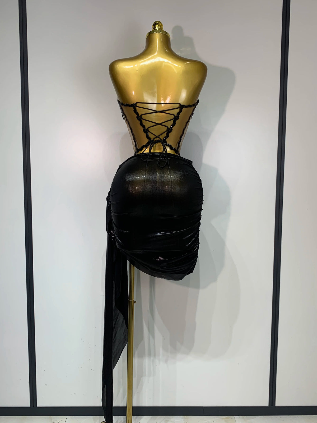 A black, ruched dress is displayed on a gold mannequin, featuring a laced-up bodice and a long black drape.