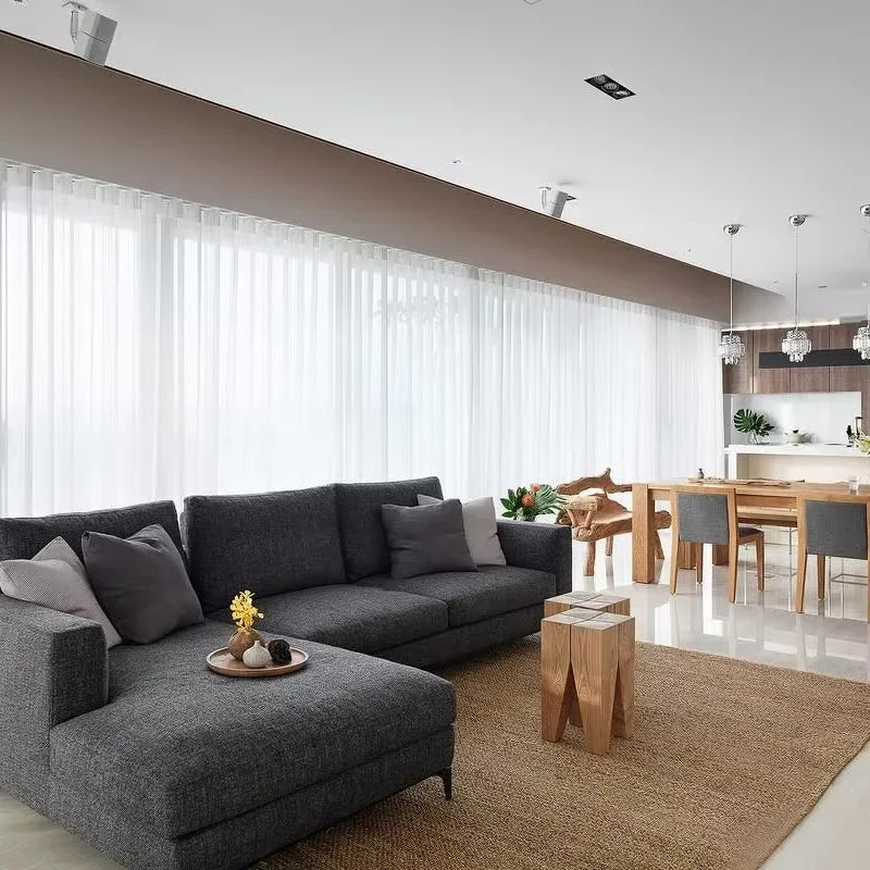 Modernes Wohnzimmer mit grauem Sofa und Holzmöbel vor durchscheinenden weißen Tüllvorhängen.