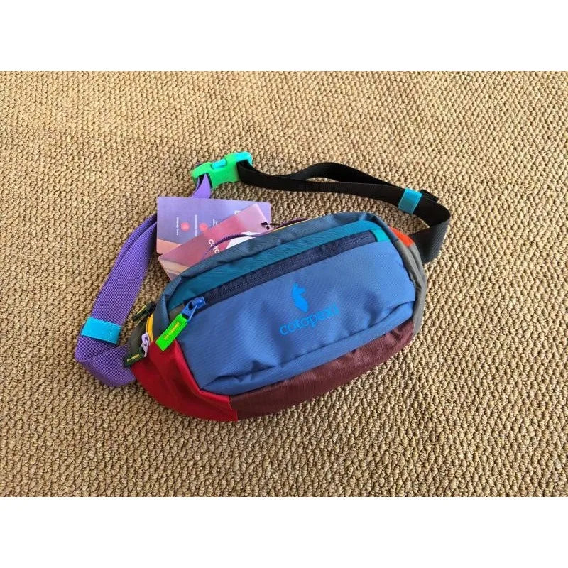 A color block waterproof nylon fanny pack in purple, red, and blue resting on a textured brown surface.