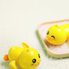 A bright yellow wind-up bath toy duck is resting on white granular material in a light pink dish.