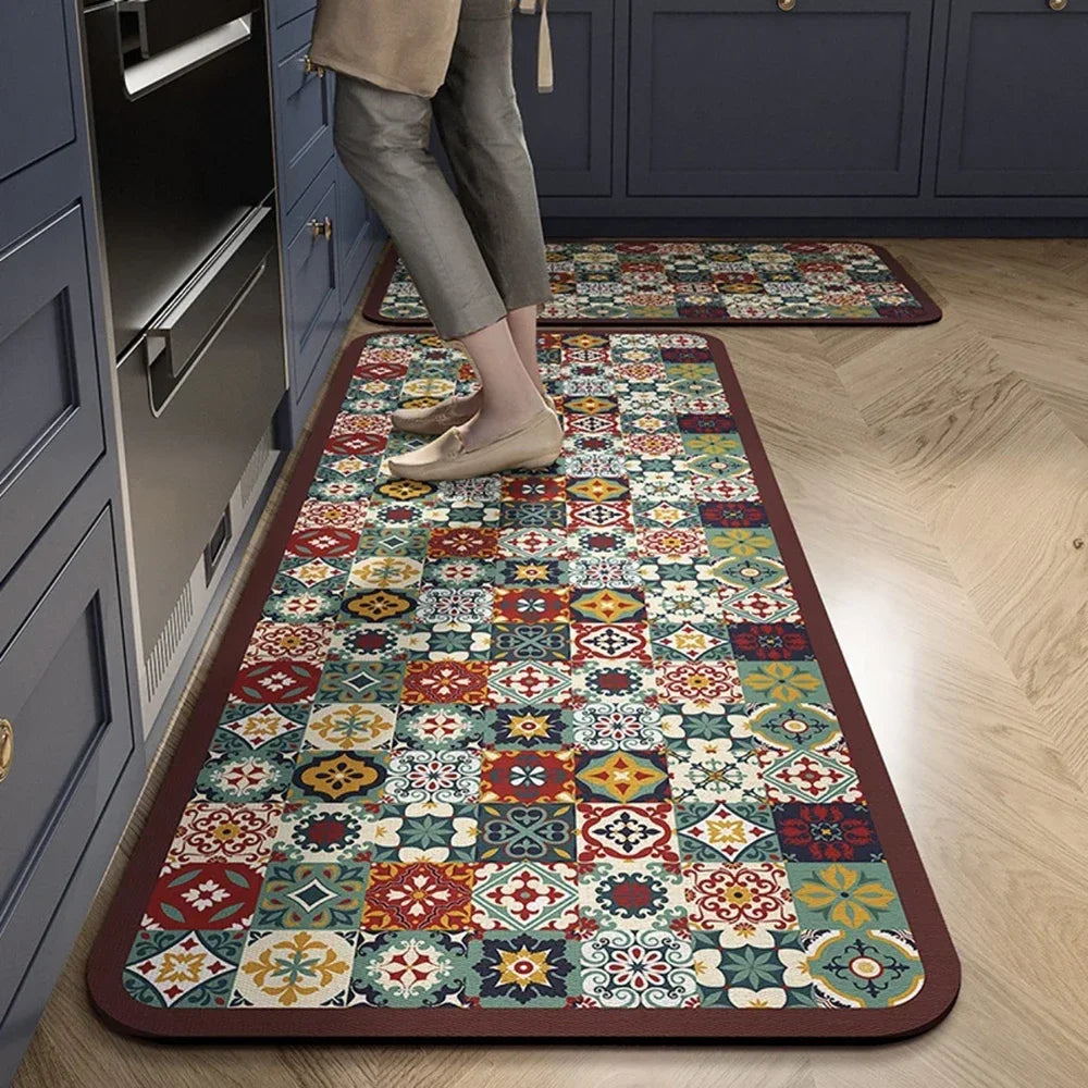A rectangular patterned floor mat with a dark border is placed on a light wood floor next to kitchen cabinetry.