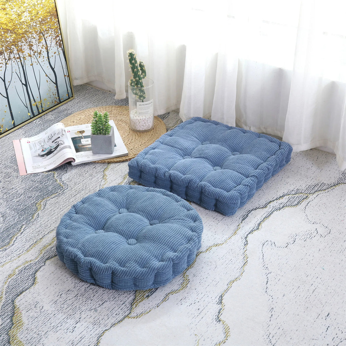 A thick blue corduroy floor cushion is placed on a light gray patterned rug next to a window.