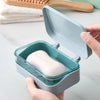 A hand holds an open, light blue transparent soap dish with a drainage tray containing a white bar of soap.