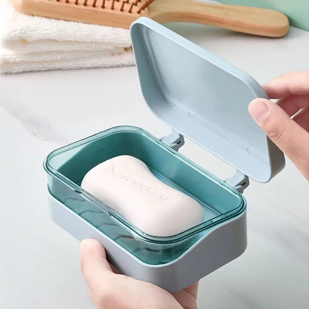 A hand holds an open, light blue transparent soap dish with a drainage tray containing a white bar of soap.