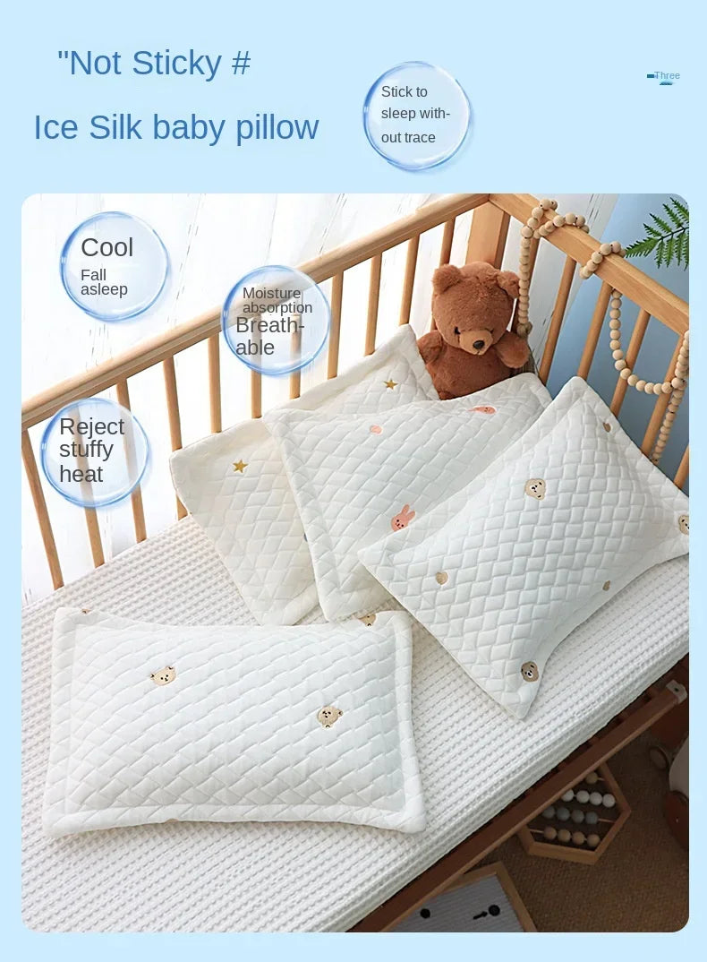 White ice silk baby pillow with a teddy bear and a small quilted pattern resting on a wooden crib railing.