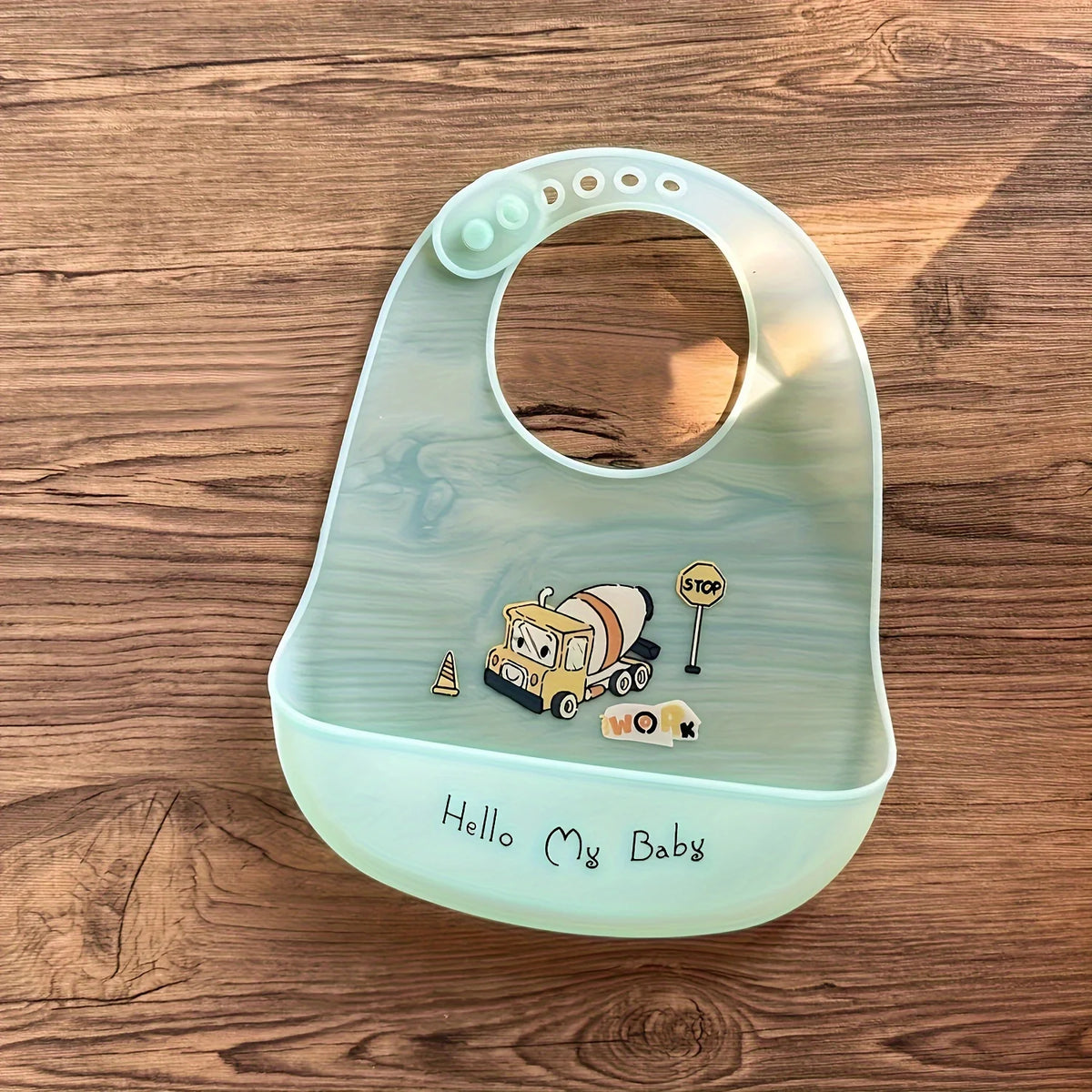A light green silicone baby bib featuring a cartoon yellow truck and stop sign on a wooden surface.
