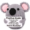 A gray crochet koala plush toy with pink inner ears and a card that reads Positive Koala