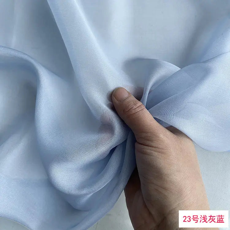A hand is folding a piece of light blue silk chiffon fabric to show its drape and texture.