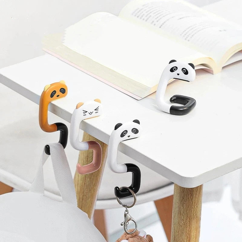 A set of cute animal-themed portable bag hooks made of silicone and plastic are displayed on a white table next to an open bo