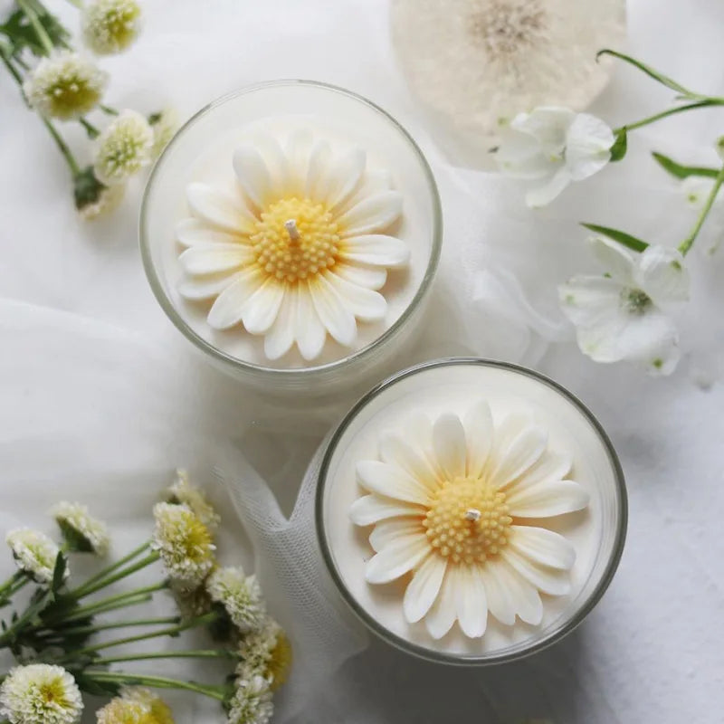 A white daisy flower is displayed in a glass, surrounded by small white flowers and greenery on a white fabric background.