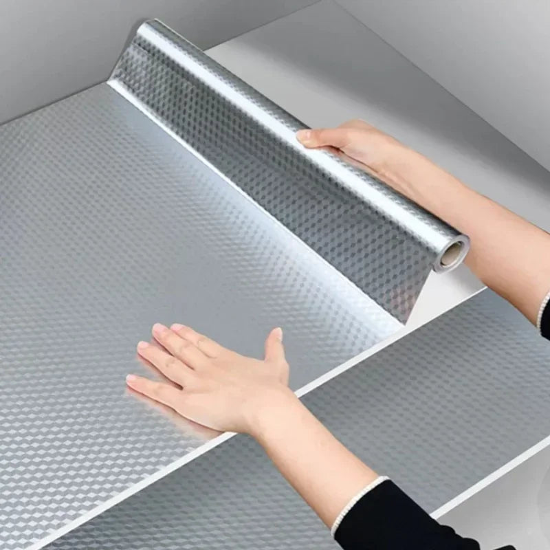 A hand is applying a roll of self-adhesive aluminum foil wallpaper to a kitchen backsplash surface.
