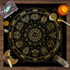 A circular gold zodiac chart with twelve signs and planetary symbols on a black background, set on a wooden surface.