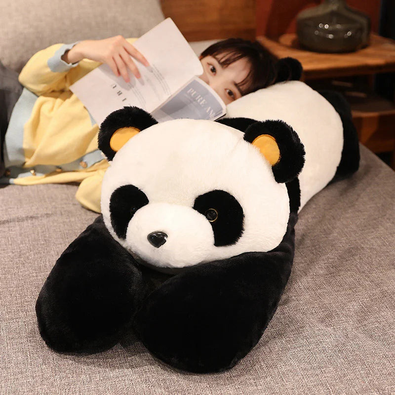A large black and white panda plush toy is resting on a gray fabric next to a person reading a book.