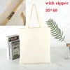 A beige canvas tote bag with a zipper is shown next to a stack of books and a fern leaf.