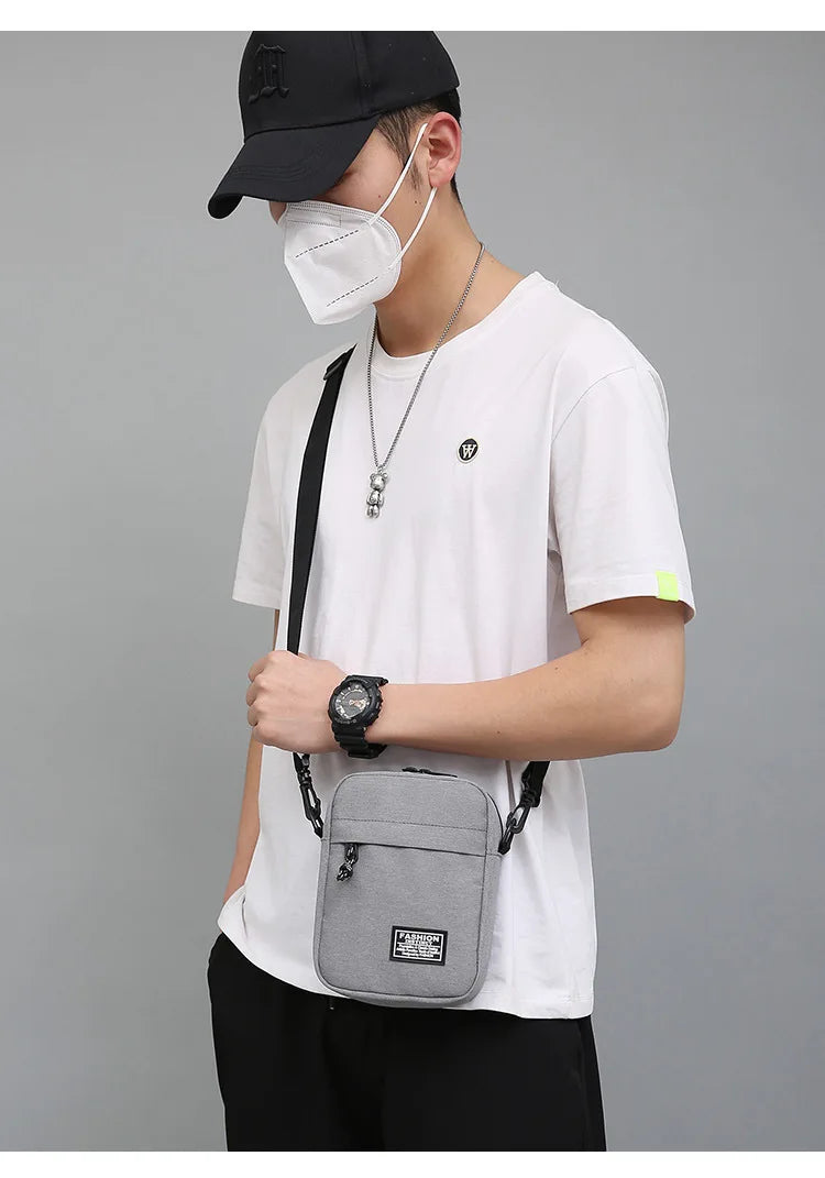 A person wearing a black cap and face mask holds a gray mini crossbody phone bag with an adjustable strap.