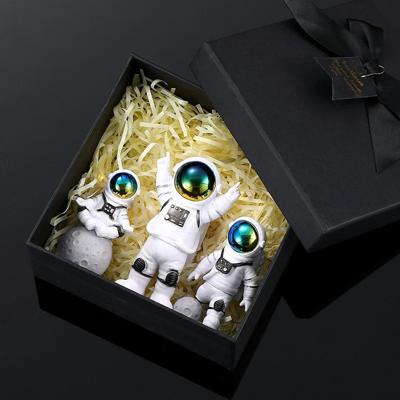 A set of three handcrafted astronaut figurines in a box with shredded yellow material and a black gift box.