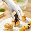A hand wearing a white disposable glove is drizzling dark syrup over a pastry on a white plate.