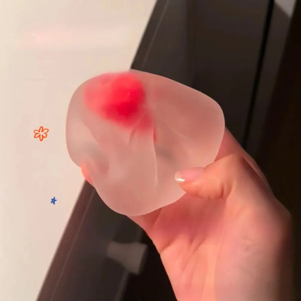 Une main serre une balle anti-stress transparente rose avec une zone rouge, démontrant sa propriété de rebond lent.