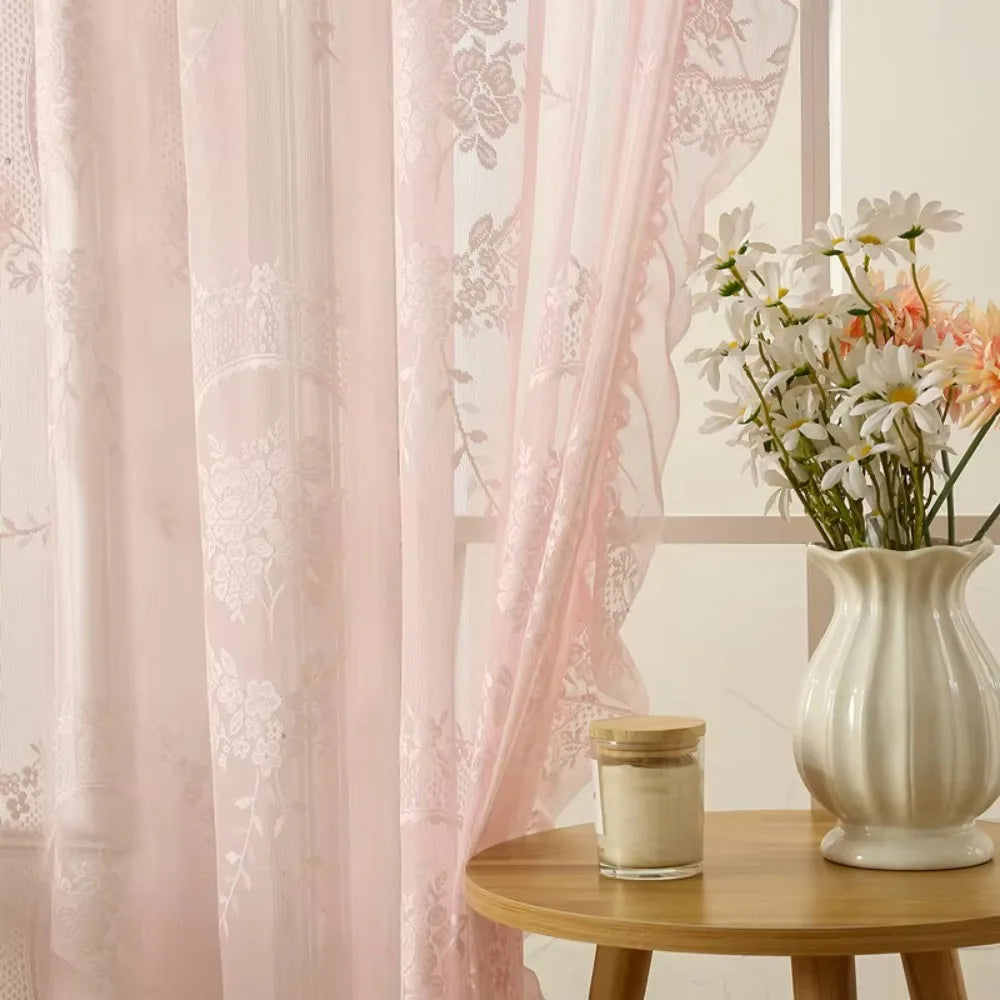 Pink sheer curtains with floral embroidery framing a vase of white and peach flowers