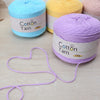 A collection of soft cotton yarn skeins in blue, yellow, and purple are displayed on a light wooden surface.
