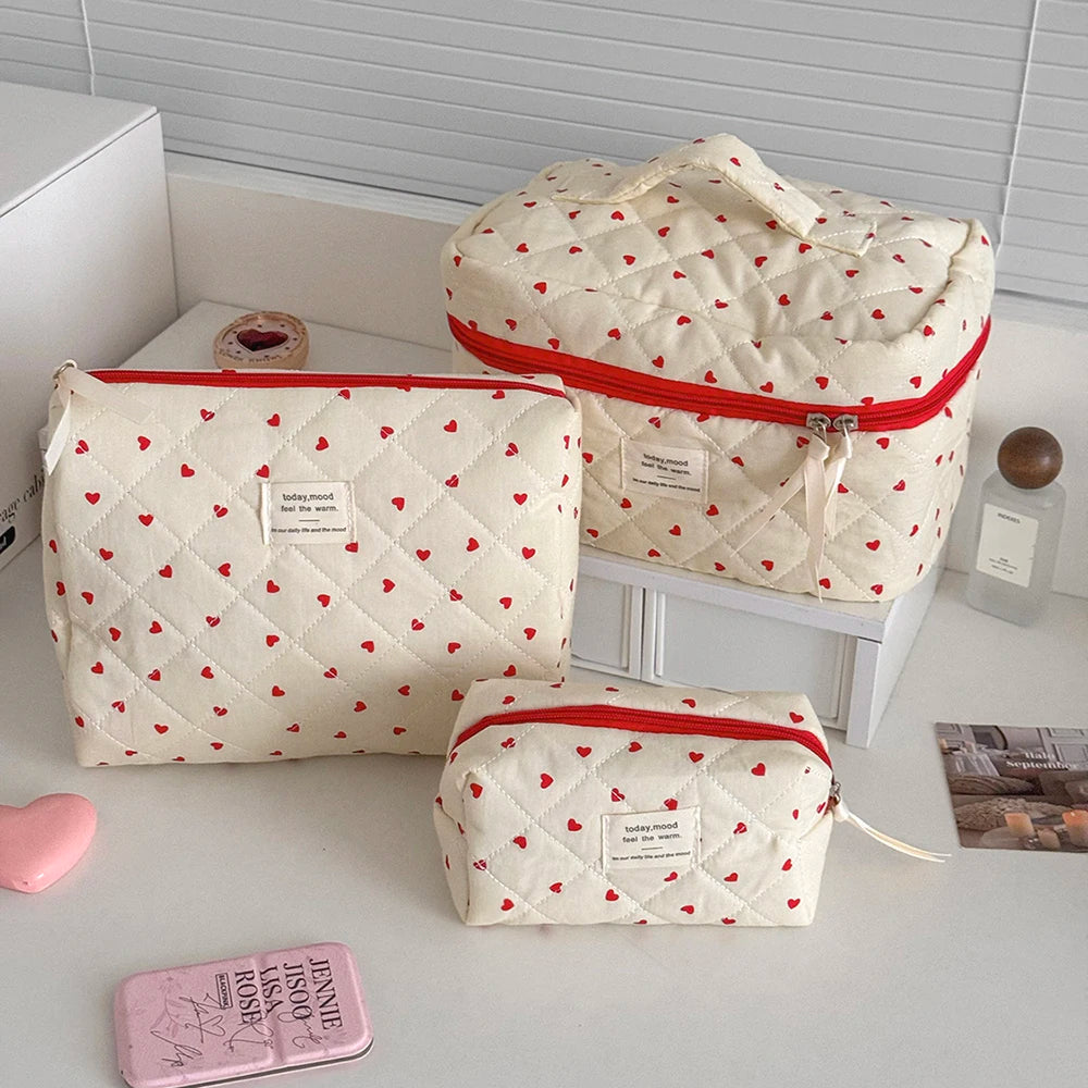 A set of quilted heart-patterned makeup bags in cream and red, including a large zippered bag and a smaller pouch.