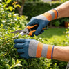 A hand wearing blue gardening gloves is using silver pruning shears on green foliage.