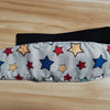 A gray fabric sleeping headrest for a baby car seat featuring colorful star shapes and a black adjustable belt strap.