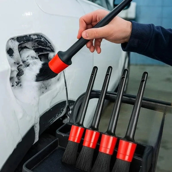 A hand uses a black cleaning brush on a white car exterior, with several red-handled detailing brushes visible in a holder.