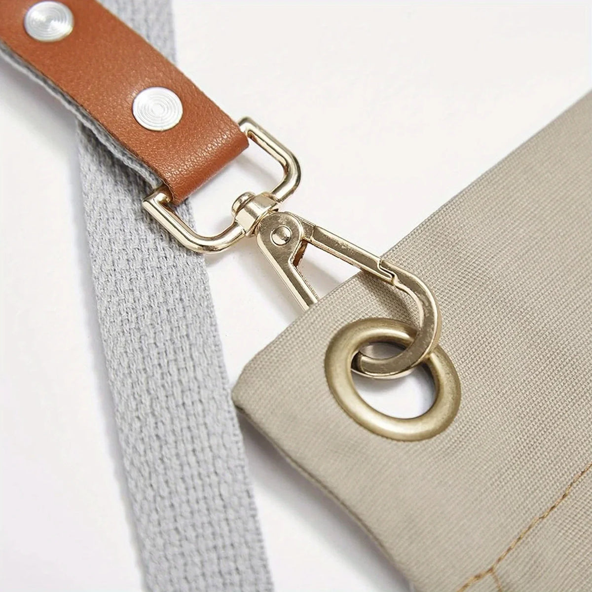 A close-up of a tan canvas apron strap with a brass hardware clip and a leather strap detail.