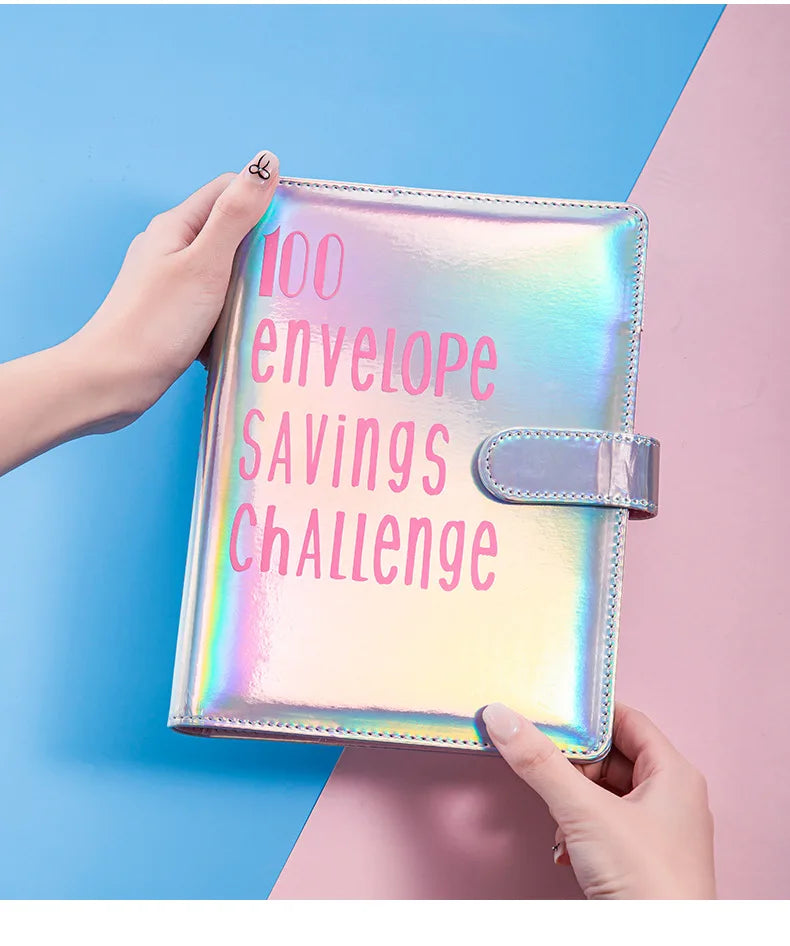 A hand holds a shiny pink envelope organizer for a 100 envelope savings challenge against a blue and pink background.