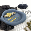 A set of gold-toned jewelry and a watch are displayed on a smooth gray silicone coaster with a black candle.
