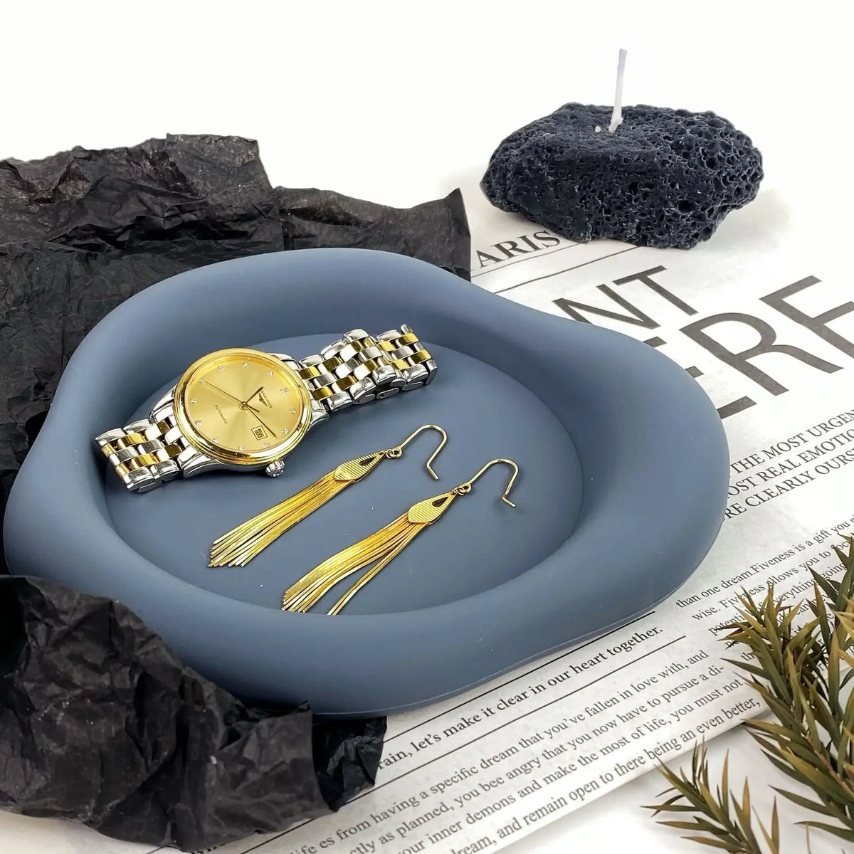 A set of gold-toned jewelry and a watch are displayed on a smooth gray silicone coaster with a black candle.