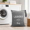 A gray fabric laundry bag with the word LAUNDRY printed on it, resting on a wooden shelf next to a white washing machine.