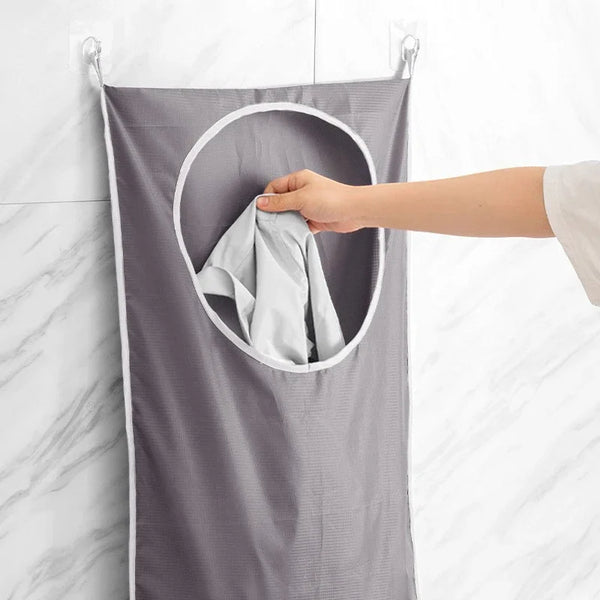 A hand is placing a folded light gray towel into the circular pocket of a gray wall-mounted laundry storage bag.