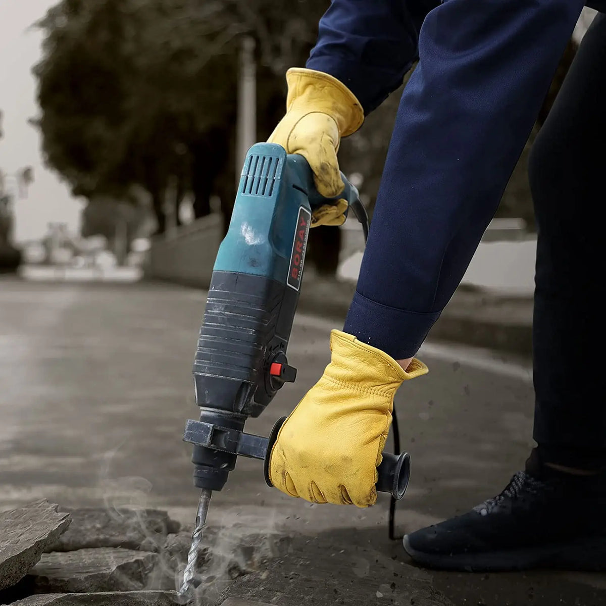 A person wearing yellow leather gloves is using a blue and black tool to prune or cut something on the ground.