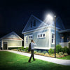 A person stands outside a house at night, looking up at a bright solar street light illuminating the front of the building.