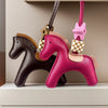 A pink leather pony charm with a gold-colored tag and a checkered yellow charm hanging from a brown leather strap.