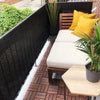 A wooden table and cushions are set on a balcony with a black privacy screen and green foliage.