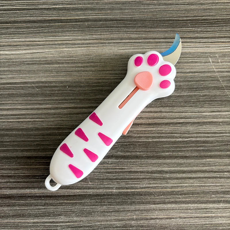 A white retractable cat paw utility knife with pink accents is shown on a wooden surface.