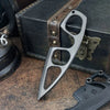 A compact stainless steel neck knife with a distinctive blade design resting on a wooden box and a dark surface.