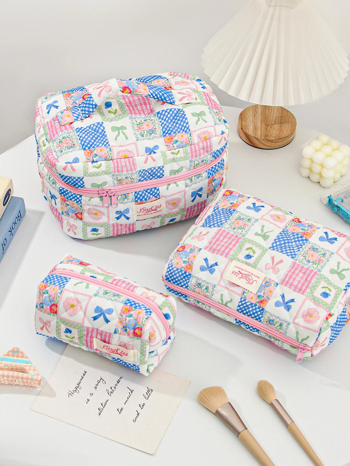 A pink and blue patchwork quilted makeup bag resting on a white surface next to a wooden object and makeup brushes.
