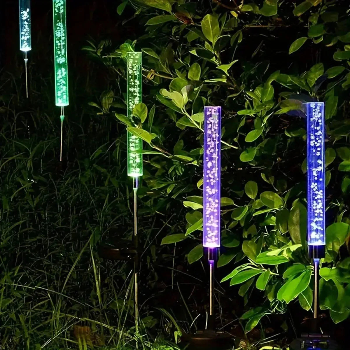 Vertical LED path lights with color-changing bubbles illuminate lush green foliage in a dark garden.