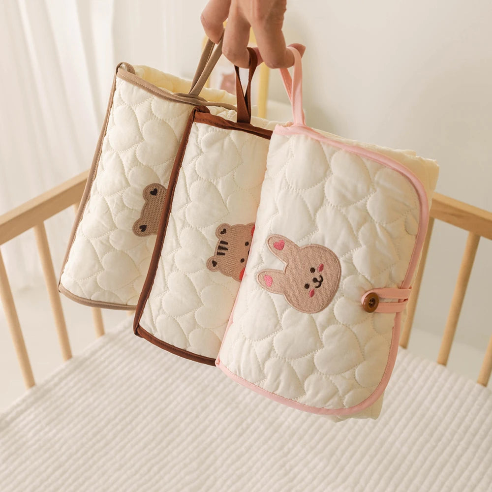 Three quilted beige and pink foldable portable diaper changing pads being held by a hand.