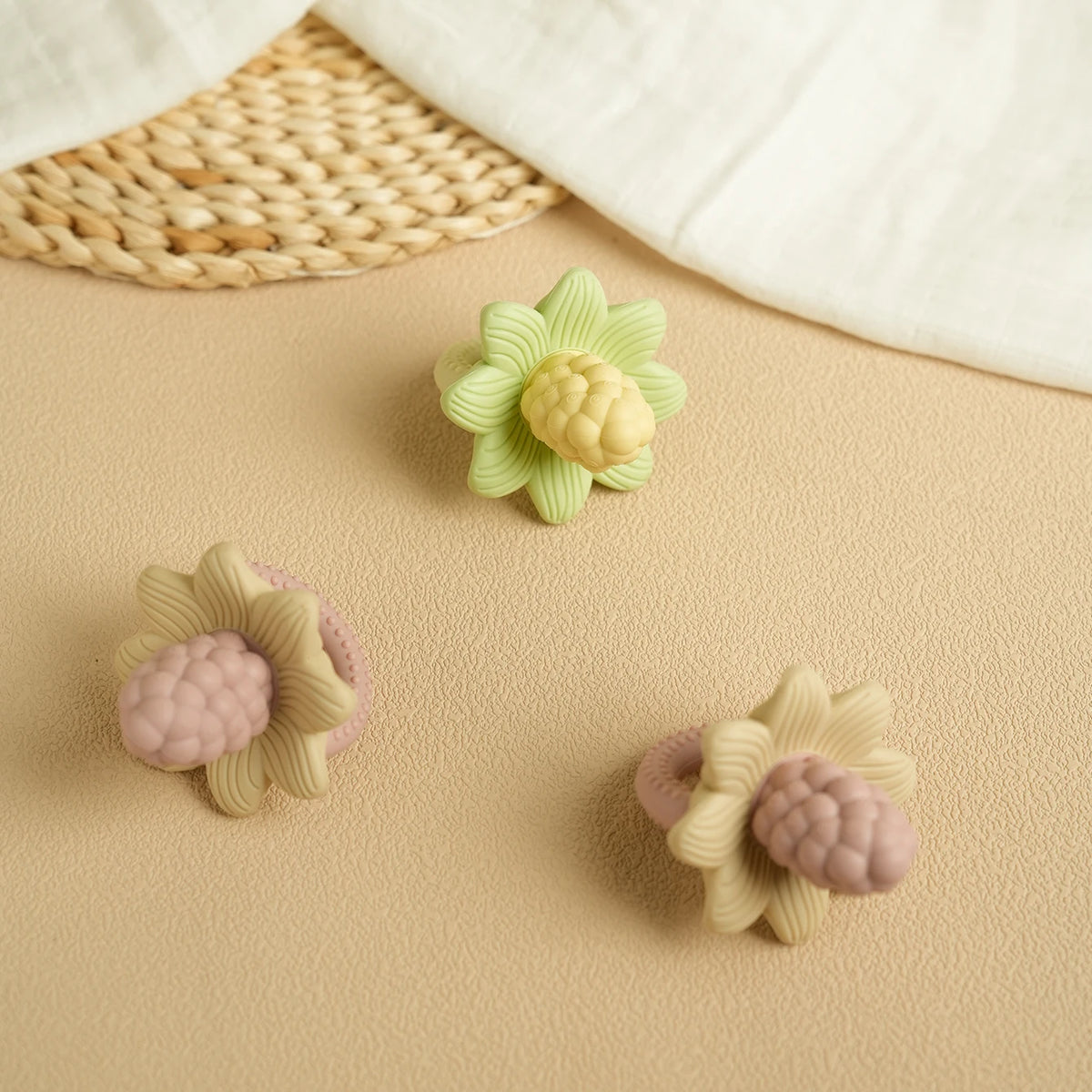 Silicone baby teether toys shaped like flowers and grapes in pale green and pink colors on a beige surface.