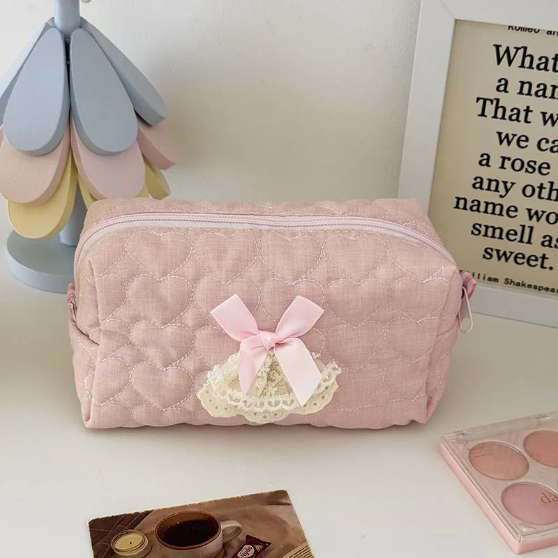 A pink quilted cosmetic pouch with a sheer lace bow trim resting on a white surface.