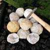 A cluster of natural agate geode crystals and light-colored stones resting on dark volcanic gravel with a wooden tool.