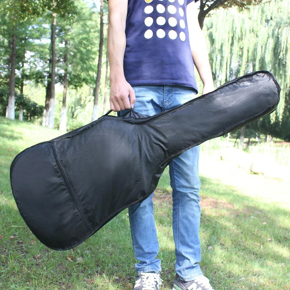 A person holds a black classical acoustic guitar soft case with a padded shoulder strap outdoors.